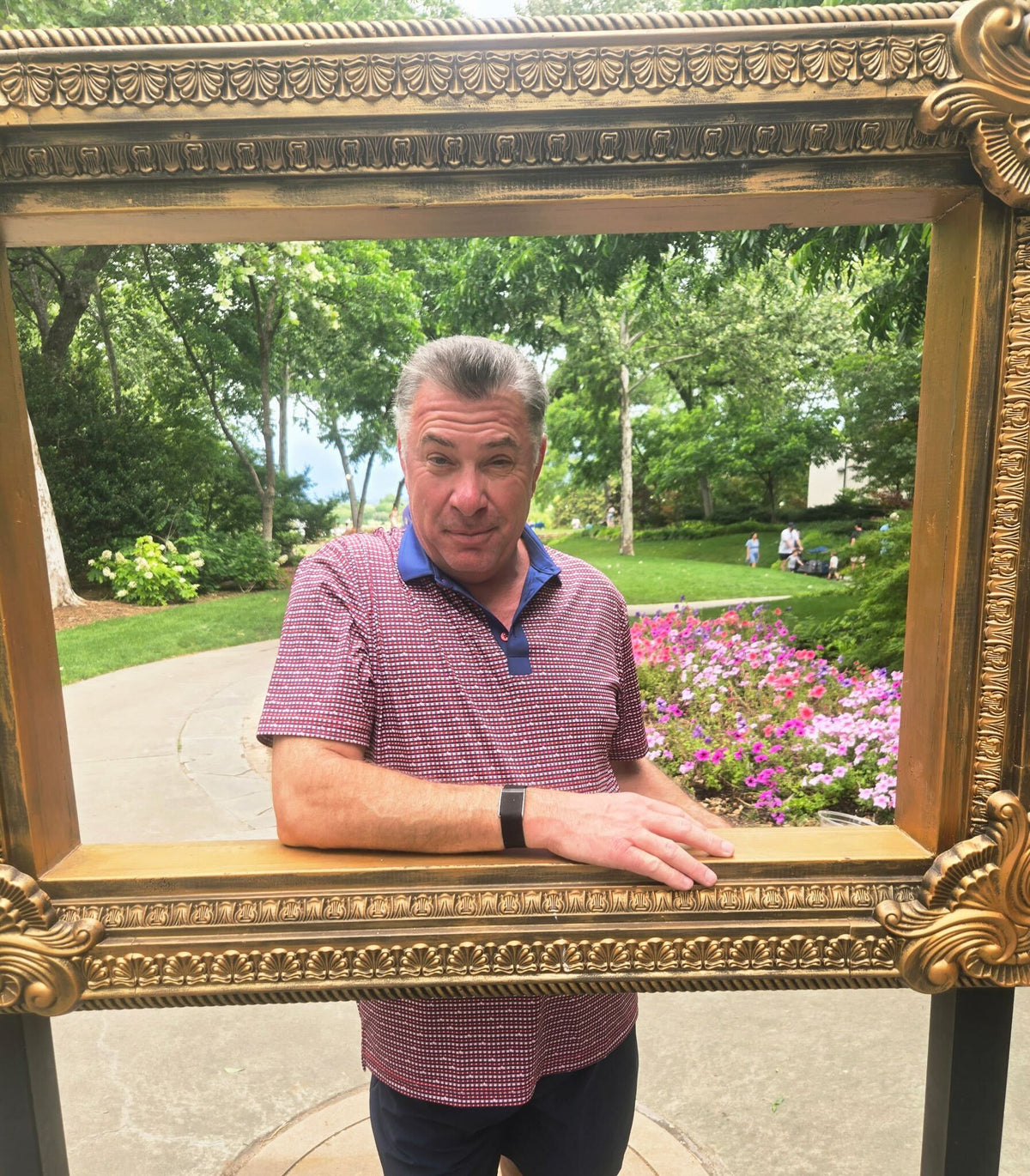 Star Spangled lifestyle — An adult wears the exact red-and-navy Star Spangled polo outdoors in a landscaped garden while posing through an ornate freestanding picture frame.