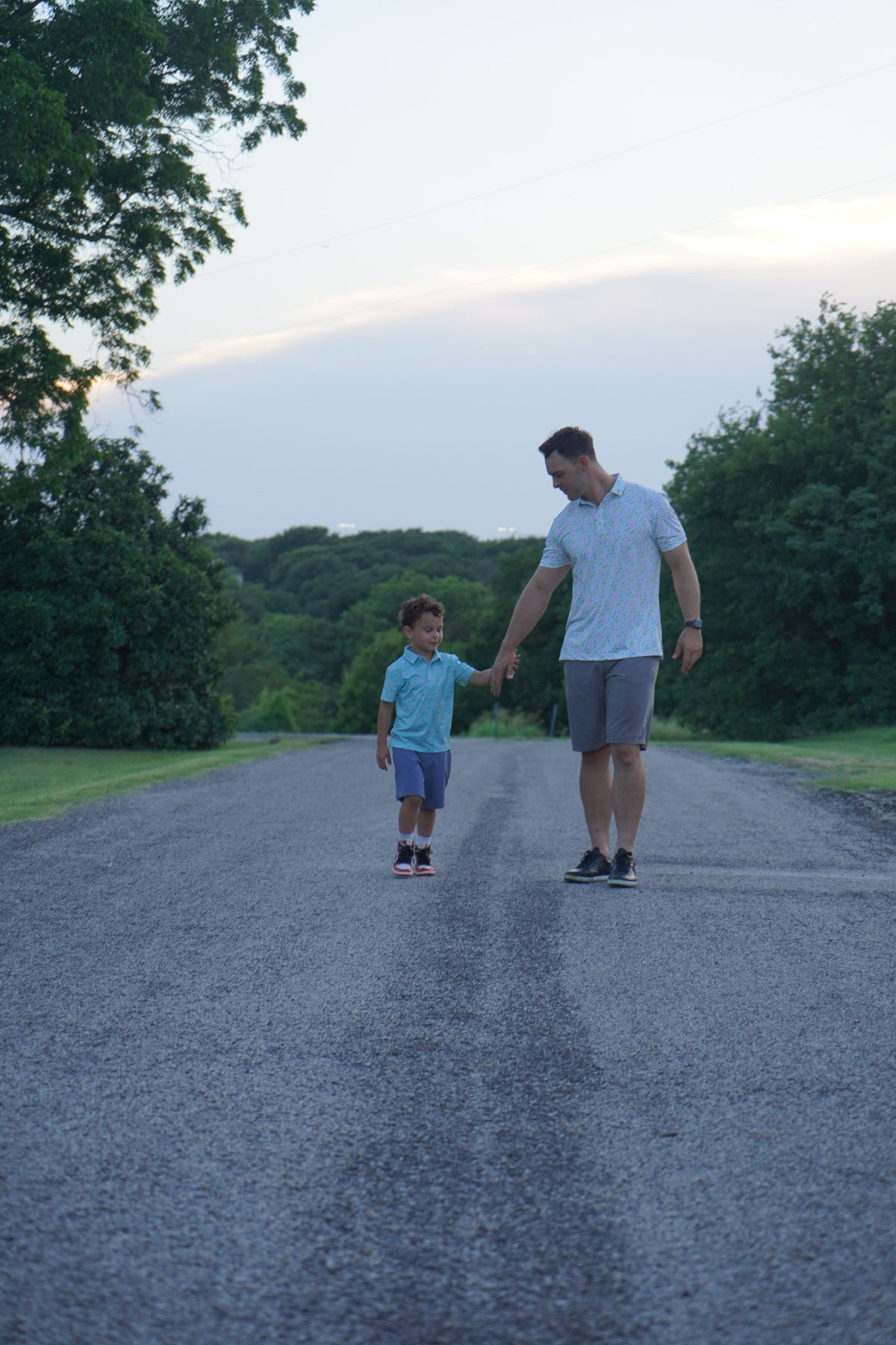 Taste of Freedom (Cream) lifestyle — A dad visibly wearing the exact cream Taste of Freedom Americana polo walks hand-in-hand with a child on a tree-lined road at dusk.